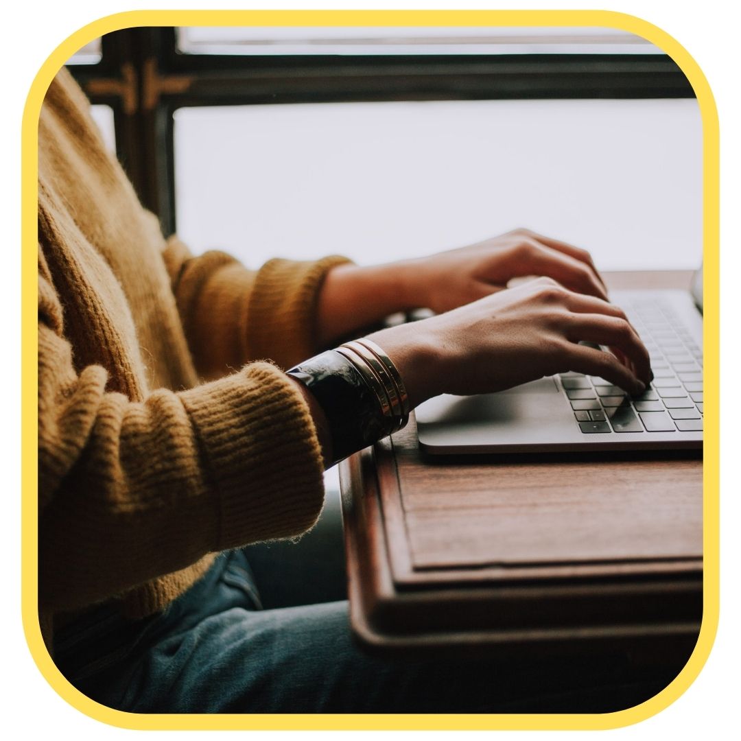 A person wearing a brown sweater types on a laptop at a wooden desk near a window, perhaps researching ways to save money on printer ink. The image has rounded corners and a yellow border.