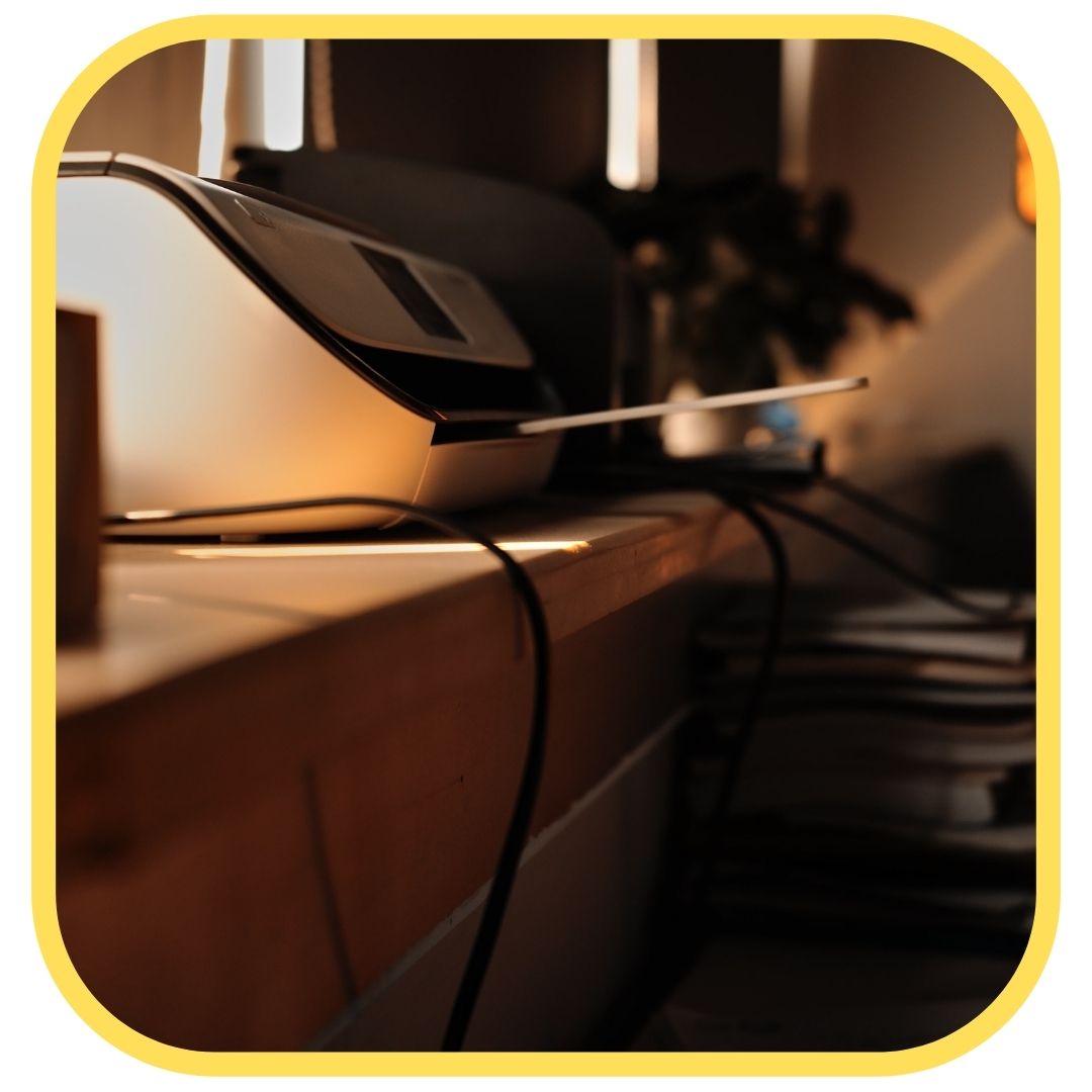A close-up of a home office setup showing a printer on a desk, with cables hanging down and soft warm lighting. The image is bordered by a thick yellow rounded frame—perfect for anyone looking to save money on printer ink.